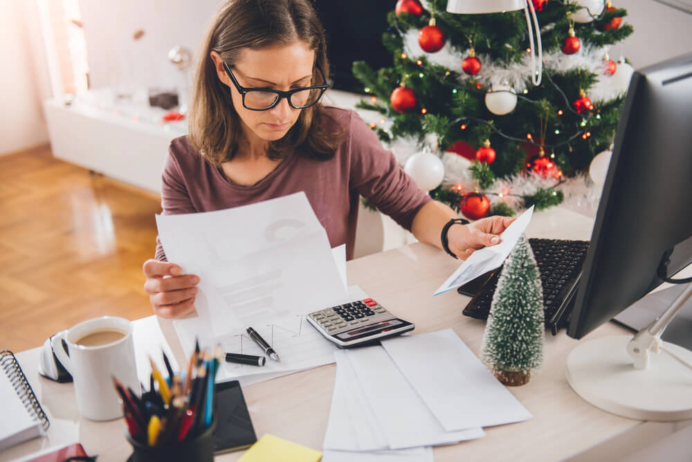 Frau liest Unterlagen an einem Schreibtisch mit Computer; im Hintergrund steht ein geschmückter Weihnachtsbaum.